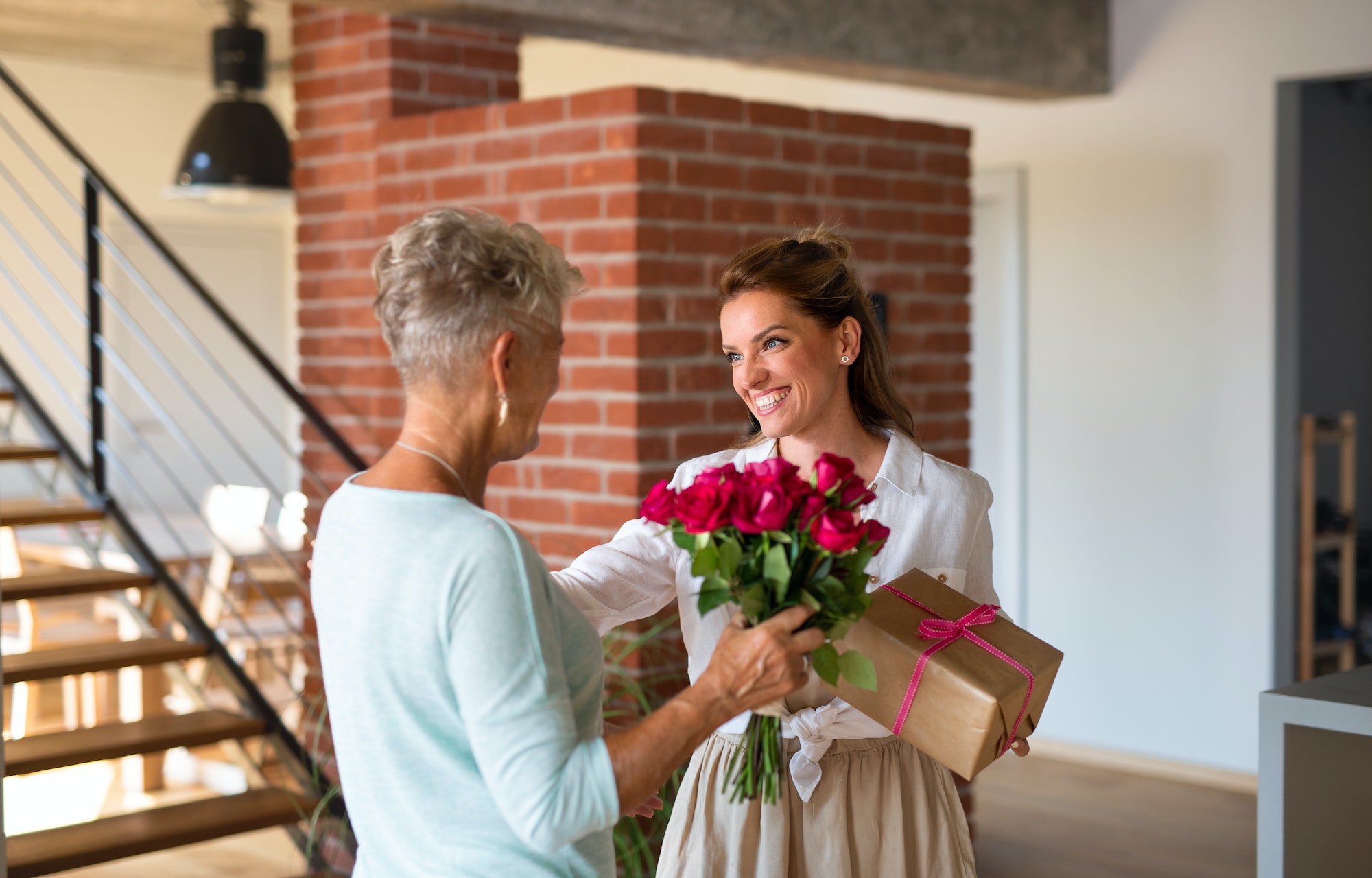 Happy adult daughter bringing gift and bouquet to senior mother indoors at home